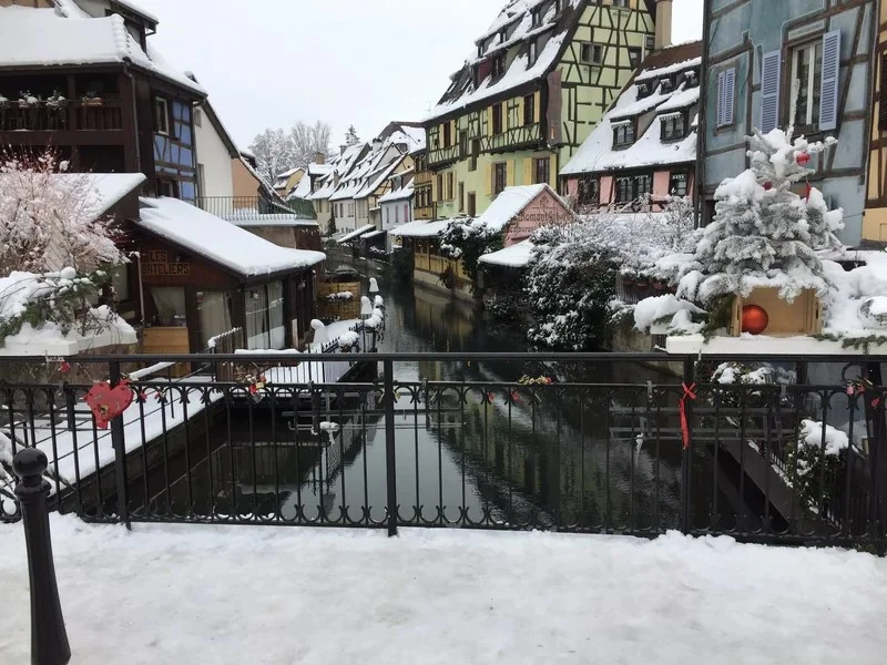 Vue du pont sur la ville de Colmar enneigée pendant les fêtes, avec maisons à colombages décorées et lumières de Noël.